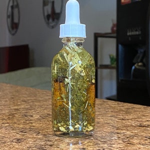 May include: A clear glass bottle with a white dropper top filled with a light yellow liquid and dried herbs. The bottle is sitting on a brown countertop.