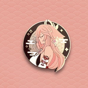May include: A gold and pink enamel pin featuring a character with long pink hair, wearing a white and gold outfit. The character is set against a black background with gold accents and a moon.