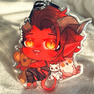 May include: A red devil character keychain with glowing orange eyes, holding a teddy bear. The keychain also features a white cat and a pink owl. The character has brown hair and horns, and is wearing black and red clothing.
