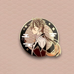 May include: A gold-toned enamel pin featuring a character with long dark hair, wearing a black and white outfit, against a black background with gold accents. The character is surrounded by a gold and white wave pattern.