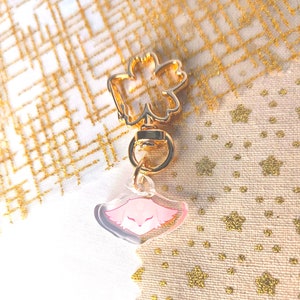 May include: A gold-colored keychain with a clear charm featuring a pink cartoon animal face. The keychain has a flower-shaped loop.