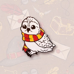 May include: A white owl with yellow and red striped scarf, enamel pin. The owl is facing the right side of the image.