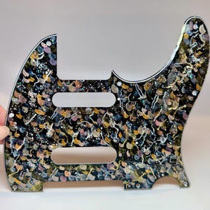 Puede incluir: Un golpeador de guitarra con un diseño abstracto y colorido. El golpeador presenta una base negra con un patrón de pinceladas doradas, rosas, azules y blancas, y purpurina. Tiene tres recortes rectangulares y agujeros para tornillos.