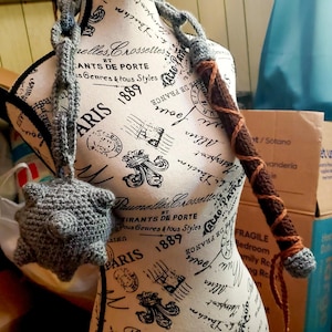 May include: A crocheted flail featuring a grey spiked ball head attached to a long grey chain and a brown handle wrapped with a brown yarn spiral. The weapon is displayed on a mannequin torso printed with black French text and decorative motifs.