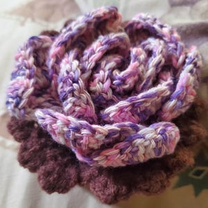 May include: A handmade crochet flower in shades of purple, pink, and white. The flower has multiple layers of petals, with a darker purple base and a lighter, variegated top. The crochet work is detailed and textured.