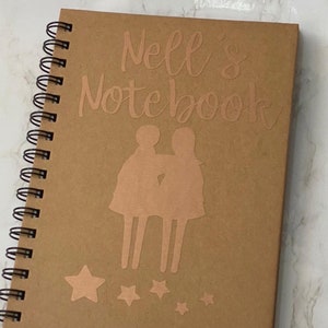 May include: A brown notebook with a spiral binding. The cover features the text "Nell's Notebook" in gold lettering and a silhouette of two people standing together. There are five gold stars at the bottom of the cover.