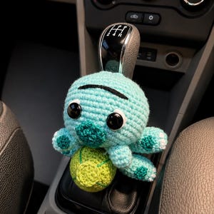 Op de afbeelding: Een lichtblauw gehaakt schildpad knuffeltje met grote zwarte ogen en een donkergroen schild, geplaatst op een versnellingspook in een auto. De schildpad heeft een klein groen balaccessoire aan de voorzijde met een textuur van garen.