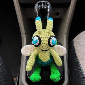 May include: A light green and dark green crocheted insect-like creature with two antennae tipped in blue, large white and blue eyes, and small white wings. The figure has a stitched black mouth and is positioned over a black car gear shift knob.