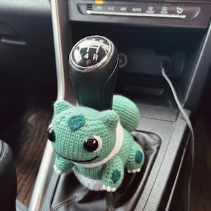 Puede incluir: Una figura de Bulbasaur verde tejida a ganchillo se encuentra en la palanca de cambios de un coche. La figura tiene ojos negros, detalles blancos y manchas verdes oscuras. La palanca de cambios es negra con letras blancas.