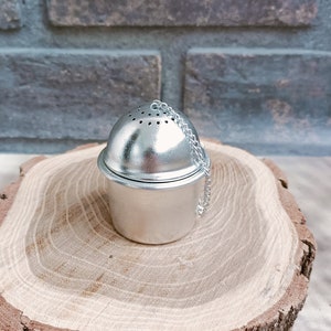 May include: Silver tea infuser with a chain, sitting on a wooden slice.