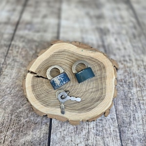 May include: Two small, vintage blue padlocks with silver metal arches rest on a wooden slice, accompanied by two keys. The locks show signs of wear, suggesting age and use. The wood slice has a natural, rustic appearance.