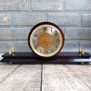 May include: A vintage wooden clock with a gold face and black hands. The clock has a brown wooden base with gold accents. The clock face reads "Vesna" and has Roman numerals.