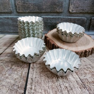May include: A set of six silver-colored metal baking cups with fluted edges. The cups are stacked on a wooden surface.