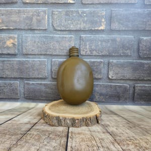 May include: An olive green canteen with a screw-on cap, resting on a natural wood slice. The canteen has a rounded, oval shape. The background features a gray brick wall and a wooden surface.