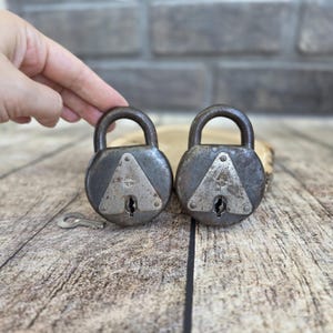 May include: Two antique, round, metal padlocks with a key. The locks are dark gray with a silver triangle design on the front. The locks have a curved handle and a keyhole. The key is silver and lies on a wooden surface.