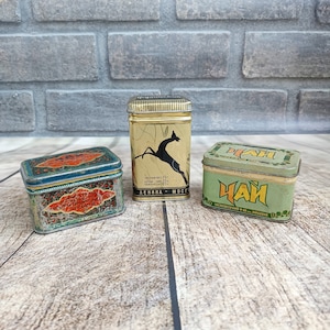 May include: Three vintage metal tins with lids. The tin on the left is blue and green with a red and orange design. The middle tin is gold with a black deer design and the text "чистый вес 75 ЦЕНА: ЧАЯ чая 22. чакницы: 21 ЛЕНИНА - МОСГ". The tin on the right is green with the text "ЧАЙ 1 ЗАТВАРИВАТЬСЯ В КИПЯЧОЙ ВОДИ".