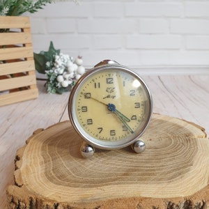 May include: A silver alarm clock with a white face and black numbers. The clock has a small bird logo and the word "Мир" written on the face. The clock is sitting on a wooden surface.