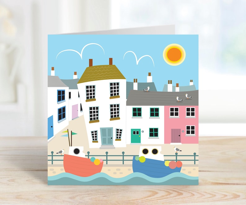 May include: A greeting card featuring a seaside town illustration. The artwork depicts colorful houses, boats, seagulls, and a bright sun against a blue sky. The card has a white border and is suitable for various occasions.