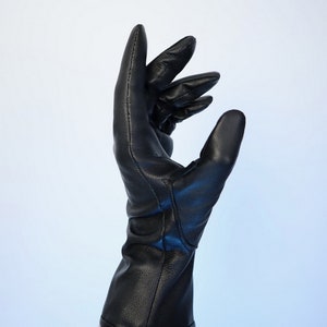 Puede incluir: Un guante de cuero negro con un pu&ntilde;o largo, usado en un brazo. El guante est&aacute; hecho de cuero liso y brillante y tiene un dise&ntilde;o cl&aacute;sico. Los dedos est&aacute;n ligeramente curvados, sugiriendo un gesto con la mano.