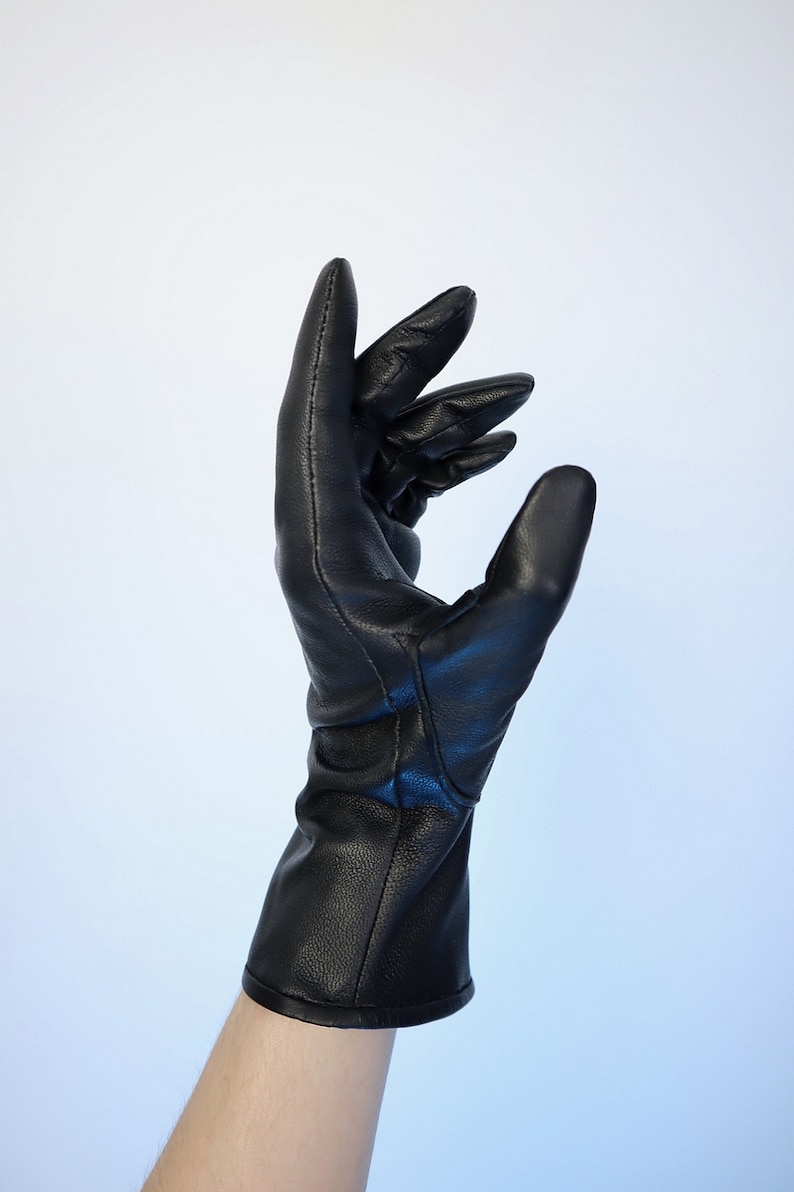 Puede incluir: Un guante de cuero negro con un pu&ntilde;o largo, usado en un brazo. El guante est&aacute; hecho de cuero liso y brillante y tiene un dise&ntilde;o cl&aacute;sico. Los dedos est&aacute;n ligeramente curvados, sugiriendo un gesto con la mano.