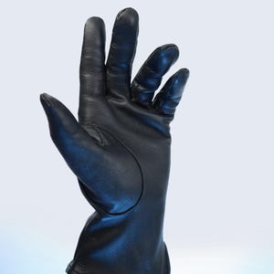 Puede incluir: Un guante de cuero negro con una textura suave. El guante se muestra con los dedos separados, mostrando el dise&ntilde;o y la artesan&iacute;a. El guante parece nuevo y sin usar, con una est&eacute;tica limpia y minimalista.