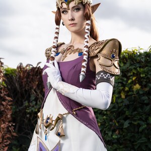 Peut inclure: Une femme v&ecirc;tue d'une robe violette et blanche avec une armure dor&eacute;e et une couronne. Elle a de longs cheveux bruns et des yeux bleus. Elle tient une &eacute;p&eacute;e dans sa main droite. L'arri&egrave;re-plan est un buisson vert.