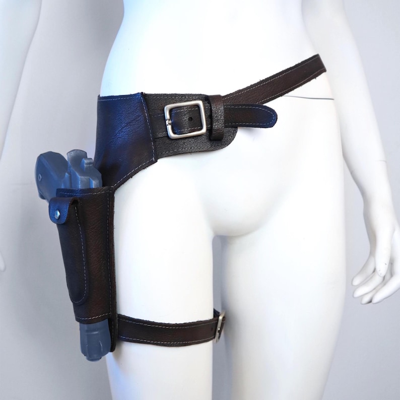 K&ouml;nnte beinhalten: Ein dunkelbraunes Lederholster- und G&uuml;rtelset. Das Holster ist f&uuml;r eine graue Pistole konzipiert und an einem breiten G&uuml;rtel mit einer silbernen Schnalle befestigt. Ein Oberschenkelriemen mit einer silbernen Schnalle ist ebenfalls enthalten.