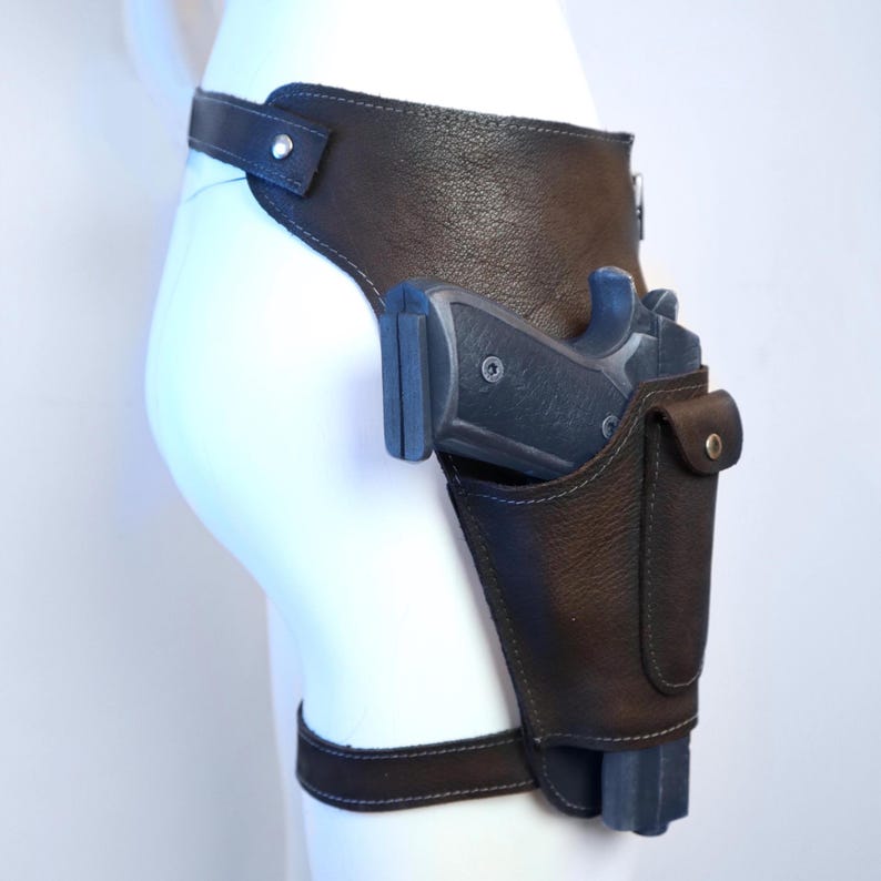 K&ouml;nnte beinhalten: Eine braune Oberschenkelholster aus Leder mit einer schwarzen Schusswaffe. Die Holster hat eine G&uuml;rtelschlaufe, einen Oberschenkelriemen und eine kleine Tasche. Die Schusswaffe ist schwarz und grau.