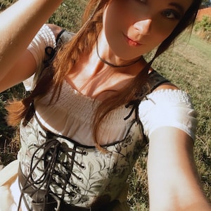 K&ouml;nnte beinhalten: Eine Frau tr&auml;gt ein wei&szlig;es Bauern-Oberteil mit einem floralen Korsett und einer schwarzen Halskette. Sie steht auf einem grasbewachsenen Feld und hat ihre Hand vor ihren Augen, als w&uuml;rde sie in die Sonne schauen.
