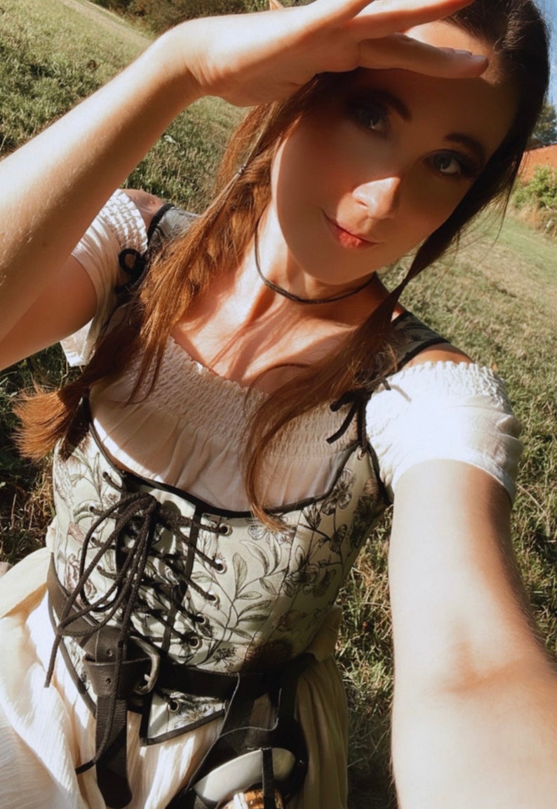 K&ouml;nnte beinhalten: Eine Frau tr&auml;gt ein wei&szlig;es Bauern-Oberteil mit einem floralen Korsett und einer schwarzen Halskette. Sie steht auf einem grasbewachsenen Feld und hat ihre Hand vor ihren Augen, als w&uuml;rde sie in die Sonne schauen.