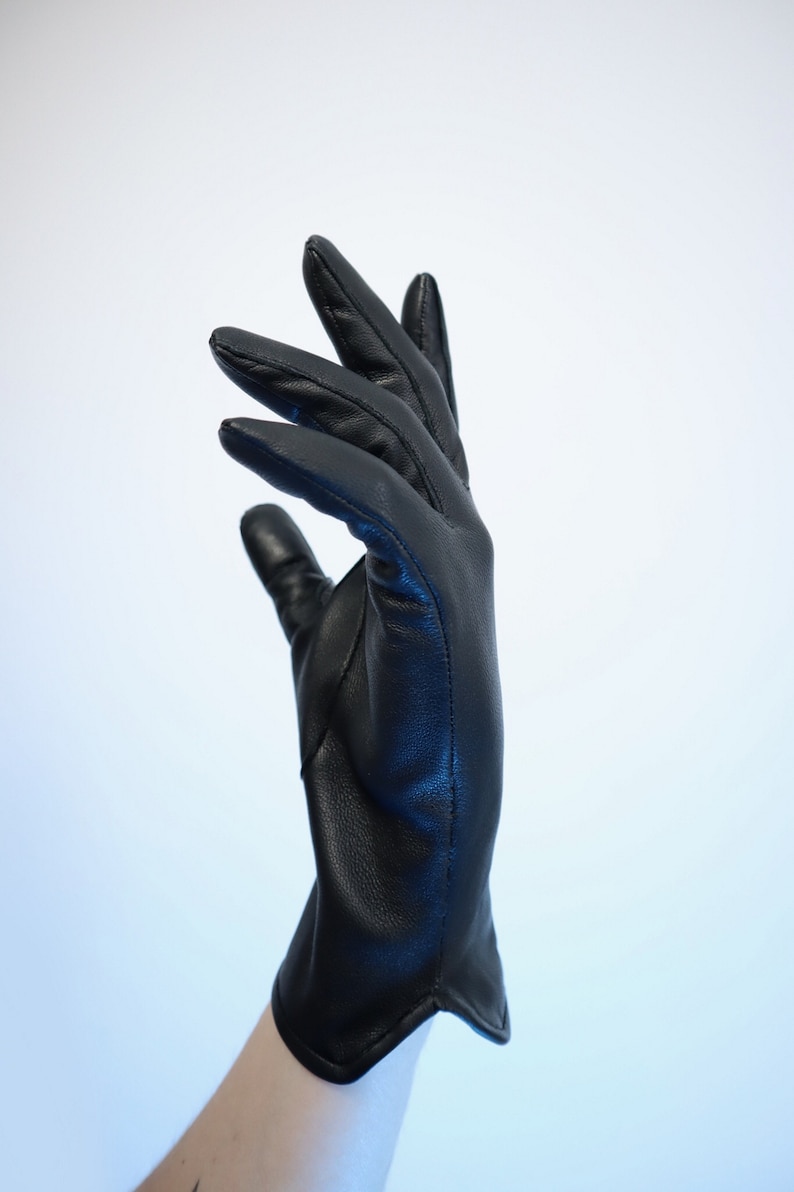 Puede incluir: Un guante de cuero negro, con costuras visibles, se muestra sobre un fondo blanco. El guante es liso y parece estar hecho de cuero de alta calidad. Los dedos est&aacute;n ligeramente curvados, lo que sugiere un ajuste natural. La est&eacute;tica general es elegante.