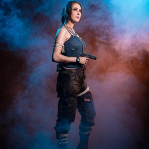 K&ouml;nnte beinhalten: Eine Frau in einem Cosplay-Outfit, die eine Handfeuerwaffe h&auml;lt. Sie tr&auml;gt ein blaues Tanktop, dunkle Cargohosen und Kampfstiefel. Der Hintergrund zeigt blauen und orangefarbenen Rauch.
