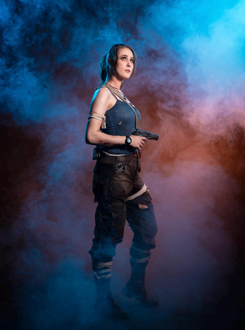 K&ouml;nnte beinhalten: Eine Frau in einem Cosplay-Outfit, die eine Handfeuerwaffe h&auml;lt. Sie tr&auml;gt ein blaues Tanktop, dunkle Cargohosen und Kampfstiefel. Der Hintergrund zeigt blauen und orangefarbenen Rauch.