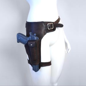 K&ouml;nnte beinhalten: Braune Leder-Oberschenkelholster mit silberner Schnalle und passendem G&uuml;rtel. Das Holster hat eine Tasche mit Druckknopfverschluss und h&auml;lt eine blaue Schusswaffe. Das Design beinhaltet einen Beingurt f&uuml;r sicheren Halt.