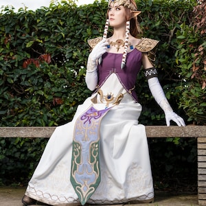 Peut inclure: Une femme v&ecirc;tue comme la princesse Zelda de la s&eacute;rie de jeux vid&eacute;o The Legend of Zelda. Elle porte une robe blanche avec un corsage violet, une ceinture dor&eacute;e et une &eacute;charpe verte et bleue. Elle porte &eacute;galement une couronne dor&eacute;e avec un bijou bleu et a de longs cheveux bruns. Elle est assise sur un banc en bois, les bras pos&eacute;s sur le dossier du banc. Elle regarde vers le haut et vers la droite.