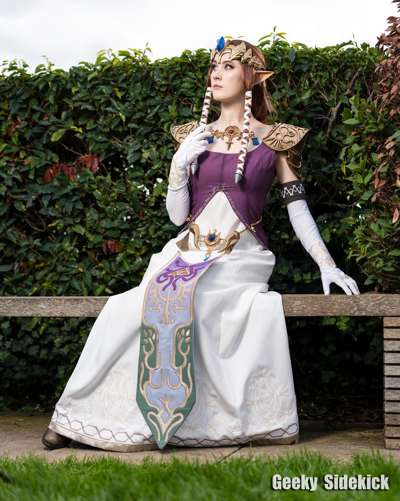 Peut inclure: Une femme v&ecirc;tue comme la princesse Zelda de la s&eacute;rie de jeux vid&eacute;o The Legend of Zelda. Elle porte une robe blanche avec un corsage violet, une ceinture dor&eacute;e et une &eacute;charpe verte et bleue. Elle porte &eacute;galement une couronne dor&eacute;e avec un bijou bleu et a de longs cheveux bruns. Elle est assise sur un banc en bois, les bras pos&eacute;s sur le dossier du banc. Elle regarde vers le haut et vers la droite.