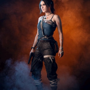 K&ouml;nnte beinhalten: Eine Frau in einem Cosplay-Outfit, bekleidet mit einem zerrissenen blauen Tanktop und dunklen Cargohosen, steht mit entschlossenem Gesichtsausdruck da. Sie h&auml;lt eine schwarze Pistole und ist von einem orange-blauen, rauchigen Hintergrund umgeben. Ihr Outfit umfasst einen G&uuml;rtel, Riemen und Bandagen.