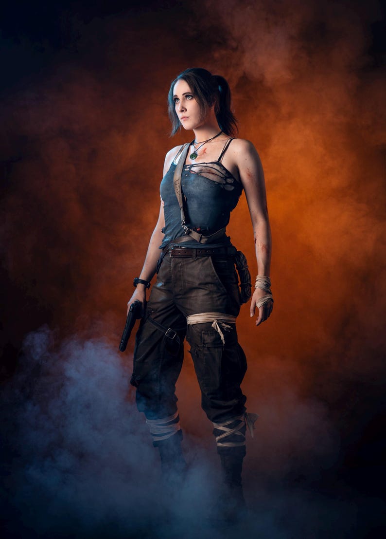 K&ouml;nnte beinhalten: Eine Frau in einem Cosplay-Outfit, bekleidet mit einem zerrissenen blauen Tanktop und dunklen Cargohosen, steht mit entschlossenem Gesichtsausdruck da. Sie h&auml;lt eine schwarze Pistole und ist von einem orange-blauen, rauchigen Hintergrund umgeben. Ihr Outfit umfasst einen G&uuml;rtel, Riemen und Bandagen.