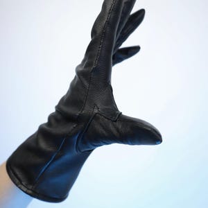 Puede incluir: Un guante de cuero negro, con una textura suave, se muestra sobre un fondo azul claro. El guante est&aacute; usado, con los dedos extendidos. La costura es visible a lo largo de las costuras. El guante parece nuevo.