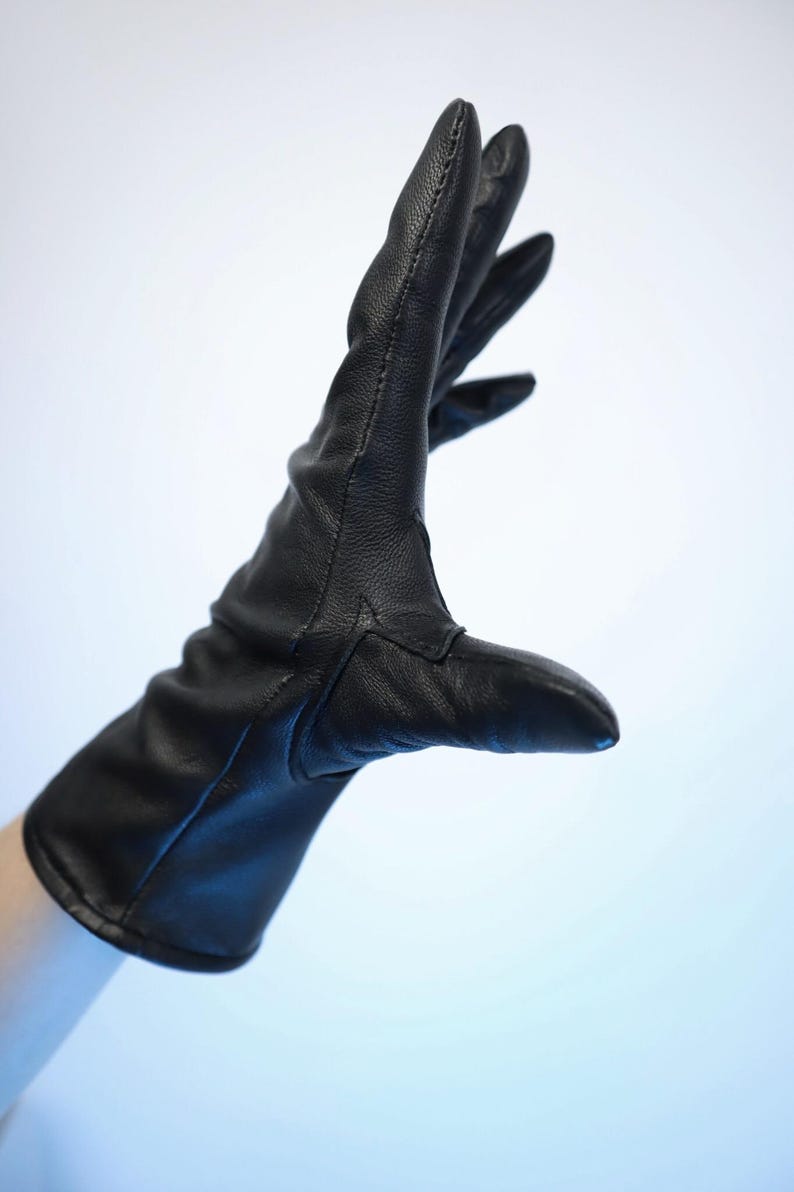 Puede incluir: Un guante de cuero negro, con una textura suave, se muestra sobre un fondo azul claro. El guante est&aacute; usado, con los dedos extendidos. La costura es visible a lo largo de las costuras. El guante parece nuevo.