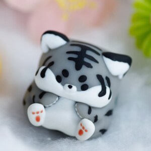 May include: A small gray and white figurine of a cat with black stripes. The cat is curled up with its paws tucked under its chin. The figurine is made of a soft, pliable material.