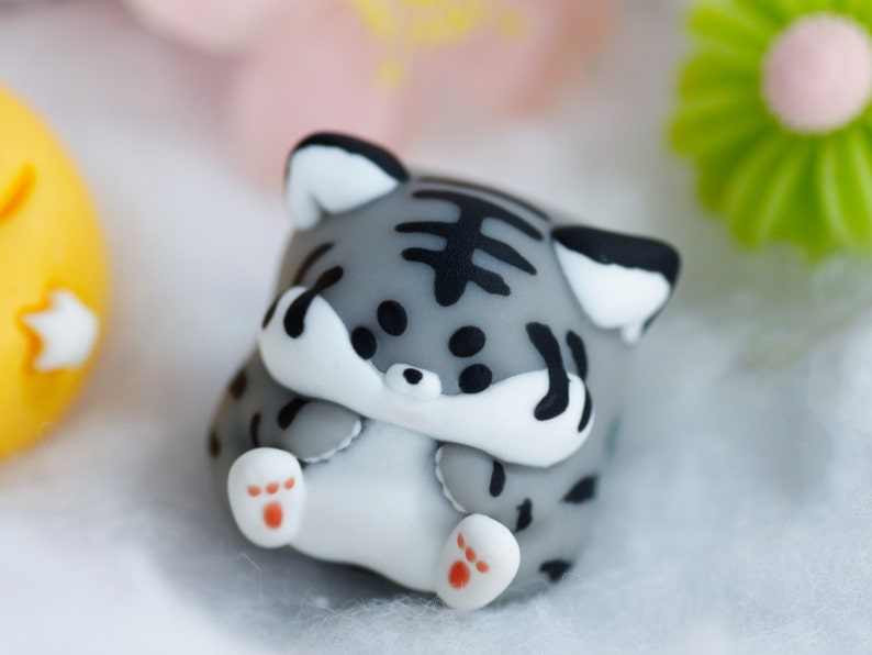 May include: A small gray and white figurine of a cat with black stripes. The cat is curled up with its paws tucked under its chin. The figurine is made of a soft, pliable material.