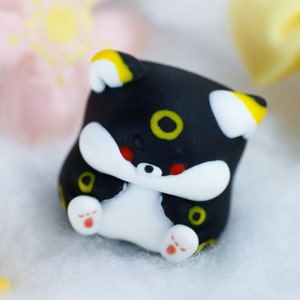 May include: A small black and white figurine of a cartoon animal with yellow accents. The figurine has a round body and a small tail. The animal has large, black eyes and a small, pink nose.