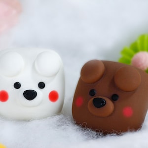 May include: Two small, square-shaped plush toys with cute animal faces. The white toy has black eyes, a black nose, and red cheeks. The brown toy has black eyes, a black nose, and red cheeks.