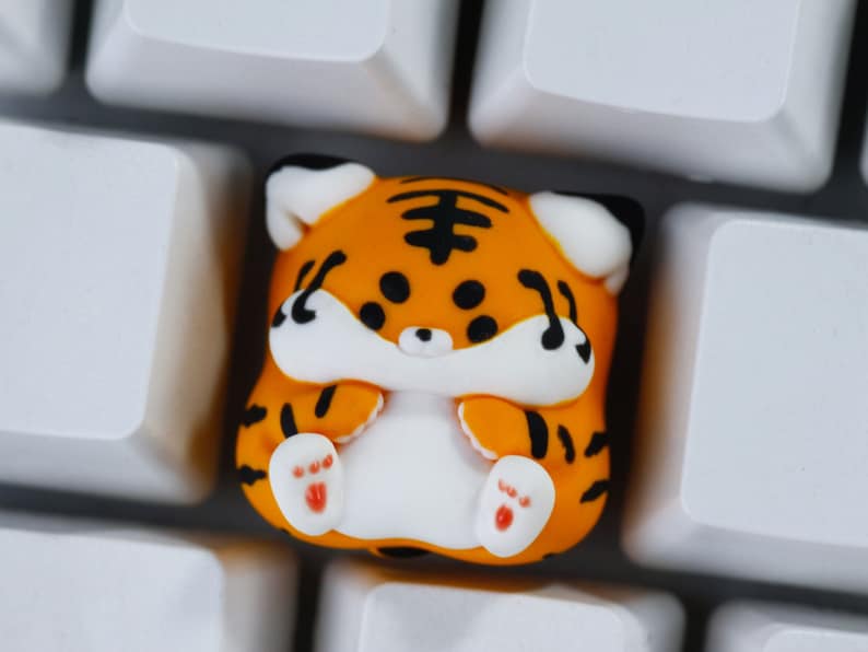 May include: A small orange and black tiger-shaped keycap with white paws and a white belly. The keycap is sitting on a white keyboard.