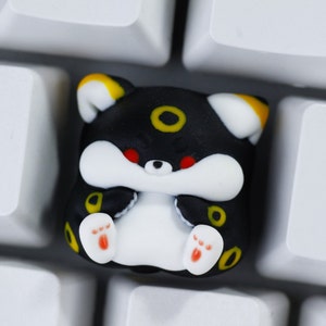 May include: Black and white keycap with a yellow and red design. The keycap features a cartoon character with a round, black eye and a white body. The character is sitting on a white keyboard.