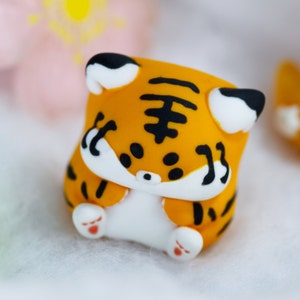May include: A small, orange and black tiger figurine with white paws and a white face. The figurine is made of clay and has a cute, cartoonish style.