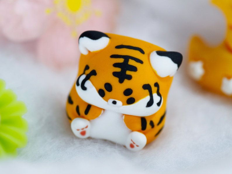 May include: A small, orange and black tiger figurine with white paws and a white face. The figurine is made of clay and has a cute, cartoonish style.