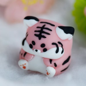 May include: A small pink and black tiger figurine with white accents. The figurine is made of fondant and has a cute, cartoonish style.