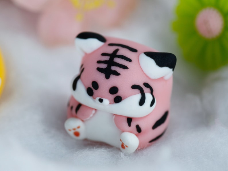 May include: A small pink and black tiger figurine with white accents. The figurine is made of fondant and has a cute, cartoonish style.
